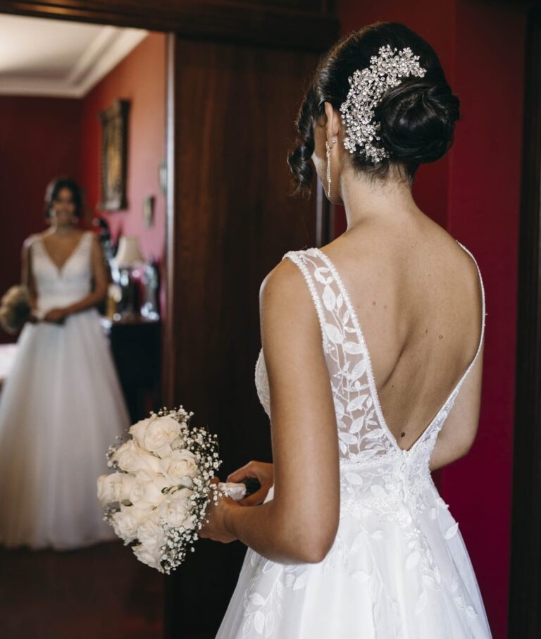 Vista de una chica de espaldas con bonito tocado y moño con adornos de strass brillante estilo floral decorativo, vestida de novia con el ramo de flores, mirandose en el espejo